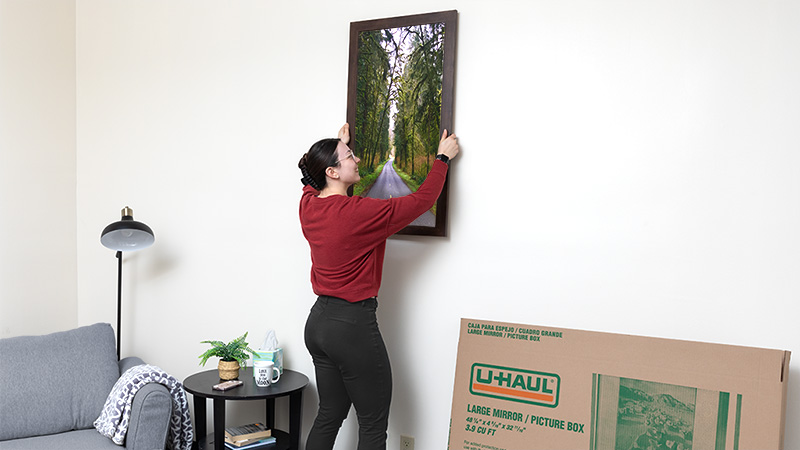 A woman considers hanging a different picture onto her wall. How to hang pictures without nails doesn’t have to be a difficult process.