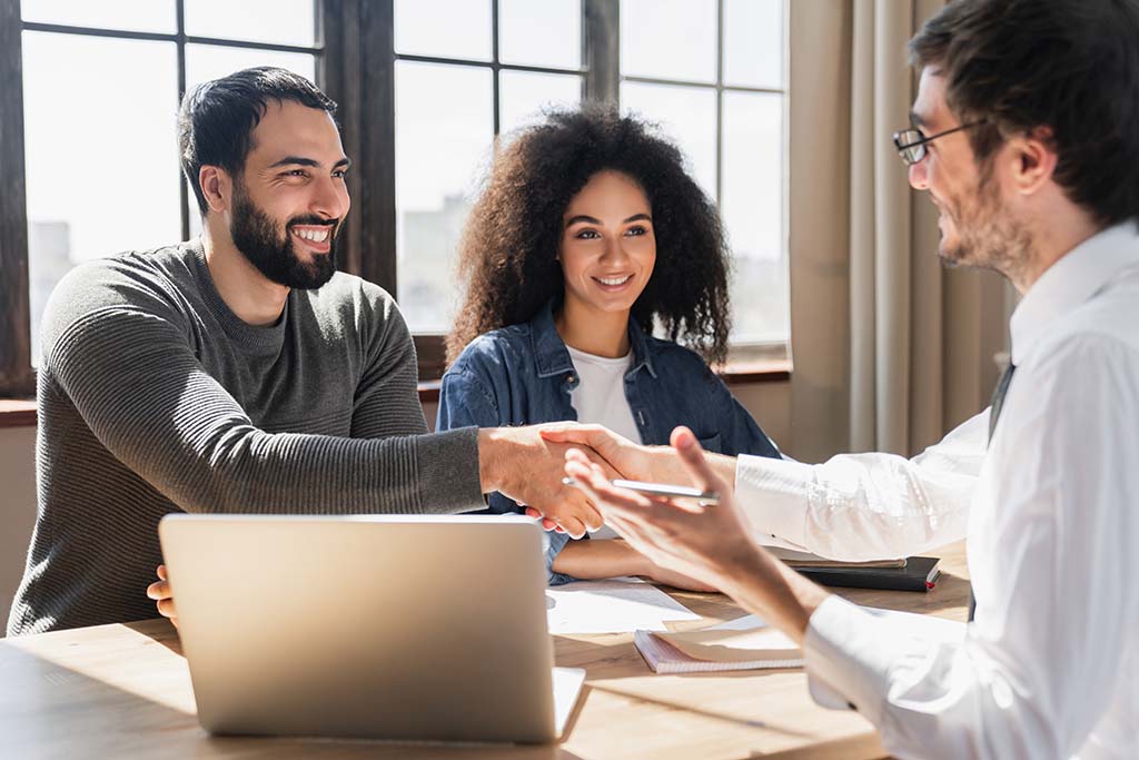A woman sits next to her husband as he shakes hands with their loan officer. The loan officer guy got the couple a good mortgage loan.