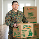 A military man stands in his empty living room while holding a small U-Haul moving box. Two moving boxes sit on the floor behind the man.