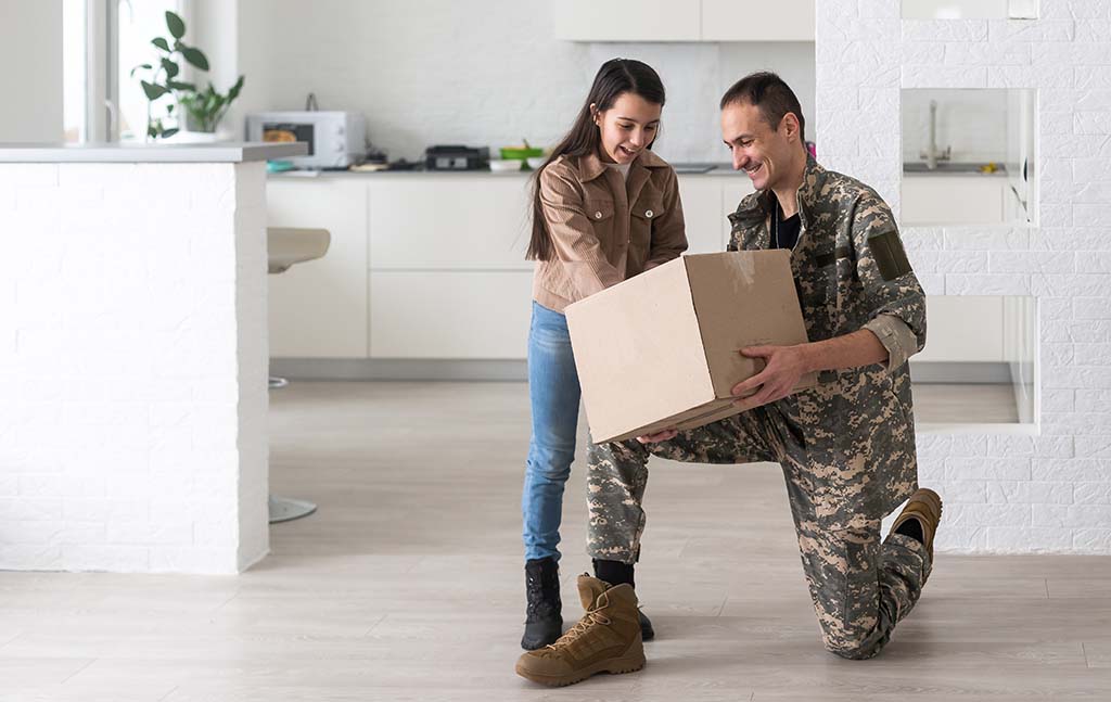 A military man kneels while showing his daughter something inside the moving box that he’s holding.