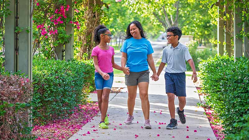 A mother holds hands with her daughter and son as they walk down a sidewalk together.
