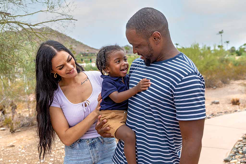 A father holds his toddler in his arm while the toddler’s mother looks at her son.
