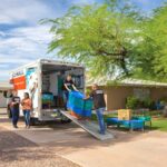 Two Moving Helpers load a customer’s piece of furniture into the back of the customer’s U-Haul truck rental. Meanwhile, another Service Provider is discussing the Payment Code with the customer to the left side of the U-Haul truck rental.