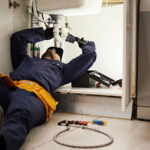 A maintenance staff member works on fixing a kitchen sink for a tenant while lying on the ground.