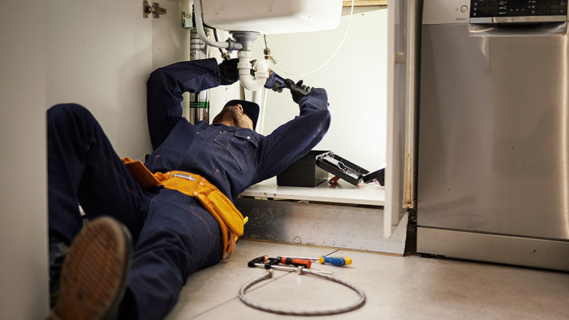 A maintenance staff member works on fixing a kitchen sink for a tenant while lying on the ground.