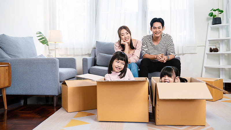 A married couple sits on the couch as their two daughters play in moving boxes in front of them in the living room of their condo.