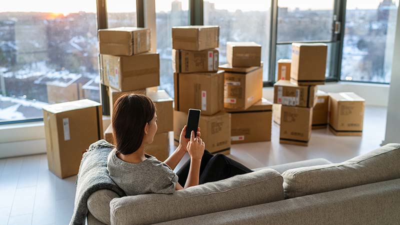 A woman sits on her couch while looking at her phone in her new condo. In front of her, multiple moving boxes rest on the floor by the window.