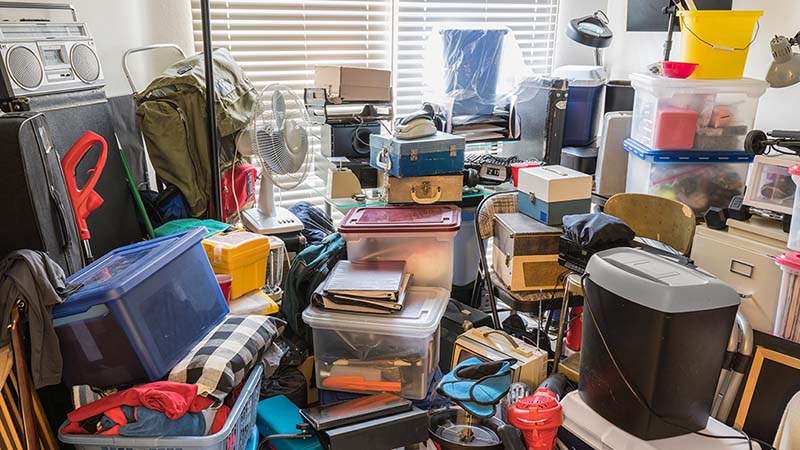 A small room is overflowing with clutter from boxes, electronics, office supplies, household objects, and miscellaneous clutter.