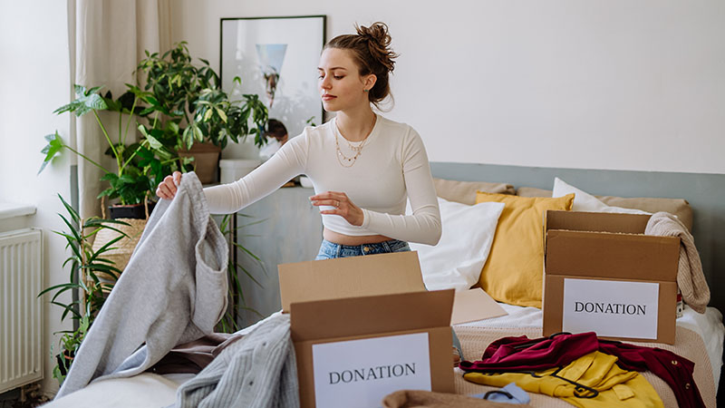 A young woman sits on her bed, sorting a pile of clothes into boxes labeled “donation.”