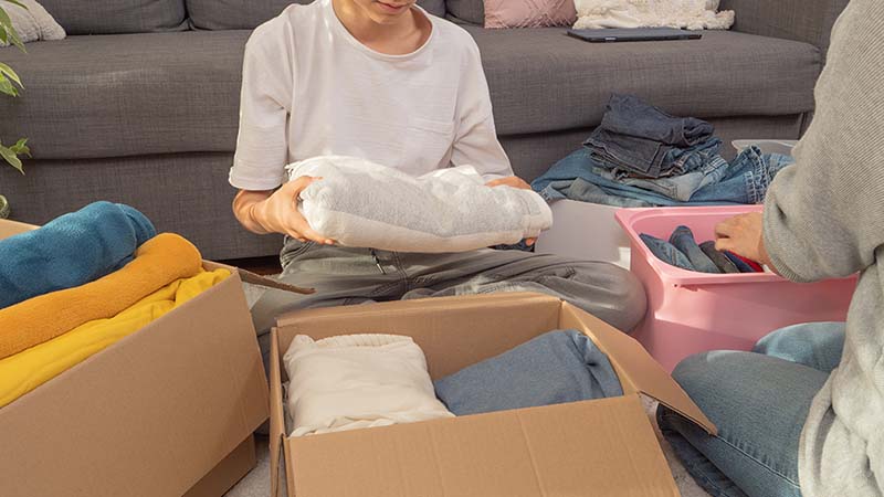Two people sit on the floor organizing clothes across two cardboard boxes and one pink plastic bin.