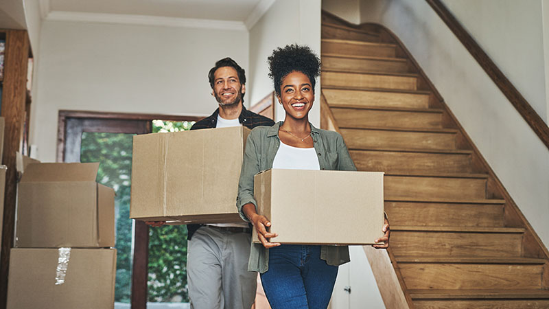 A man and a woman walk inside their new home while carrying a moving box. The couple discussed questions to ask before moving in together.