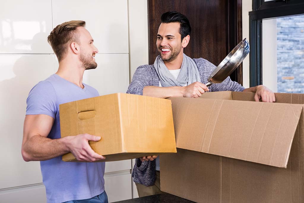 A gay man and a bisexual man smile at each other as the bisexual man pulls out a frying pan from a moving box while the gay man sets down a moving box on the countertop in their new kitchen. The gay couple discussed questions to ask before moving in together.