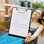 A woman shows a clipboard with a piece of paper on it. The document says “Contract” in large letters before going into the specifics of the short-term lease agreement.