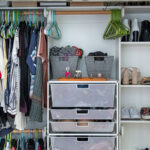 On the left side of a small closet, shirts hang from two clothing rods. In the middle, a set of mesh metal drawers is full of clothes. On the right side, shoes sit on four white shelves.