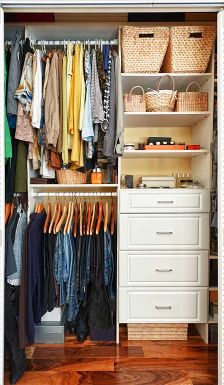 On the left side of a small closet, shirts hang from a tall rod while pants hang from a lower rod. On the right side, the closet has a set of white wooden clothing drawers. Four white shelves holding wicker baskets sit above the drawers.