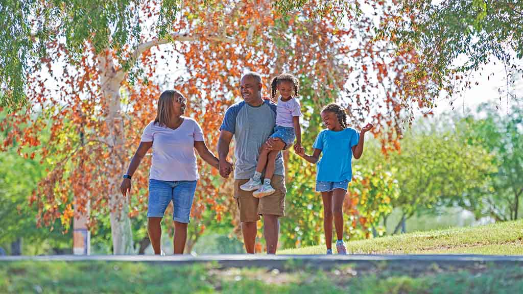 A father and mother hold hands as they walk in a park with their two daughters. The father is carrying one of the two daughters.