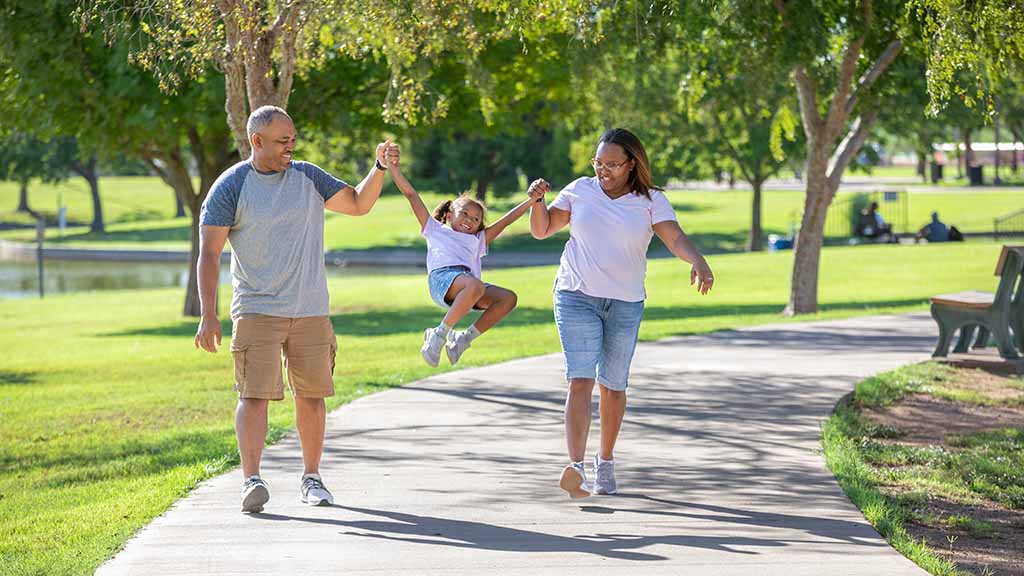A father and mother swing their daughter into the air on the sidewalk while holding their daughter’s hands in the park.