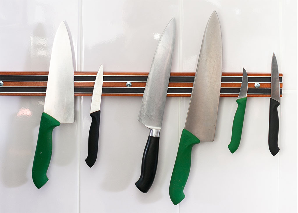 A wooden magnetic knife strip hangs on a white tiled wall of a tiny kitchen. Six knives of varying sizes adhere to the knife strip.