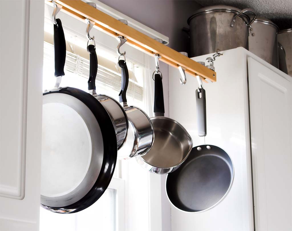 A wooden bar hangs in front of a kitchen window between two sets of cabinets. Five assorted pots and pans hang from hooks on the bar.