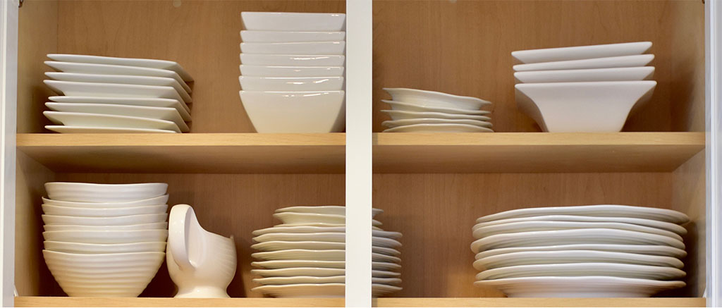 Stacks of assorted white bowls and plates sit on an open wooden cabinet in a tiny kitchen.