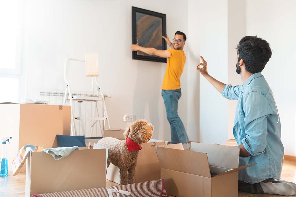 A gay man gives an OK sign to his boyfriend who's hanging a picture on the wall of their new home. Moving boxes and the gay couple’s dog surround the two men.