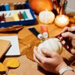A table is covered with arts and crafts supplies, including paints, paper bags, autumn leaves, pumpkins, and candles. In the foreground, a person is holding a small, white pumpkin and a marker.