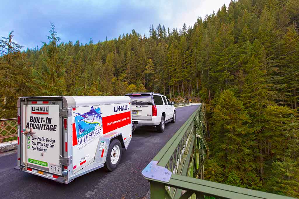 A car towing a U-Haul cargo trailer is driving on an asphalt road surrounded by pine trees on a fall afternoon.