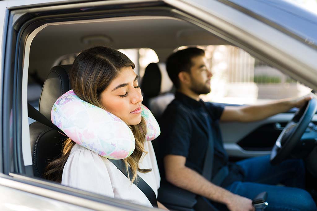 A woman sleeps using her travel neck pillow while her boyfriend drives the car on their moving road trip. Travel neck pillows are great going away gifts.