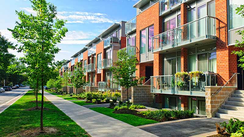 Brick townhouses line next to one another with small, private patios. A grassy area and a sidewalk lead up to the townhouses.