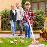 A couple looks at their daughter as she rakes some fall leaves in the family’s backyard. A bag filled with leaves sits next to the girl.