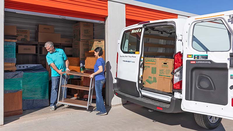 A couple carefully moves a piece of furniture into their storage unit. The couple has more U-Haul moving boxes in their U-Haul cargo van to unload.