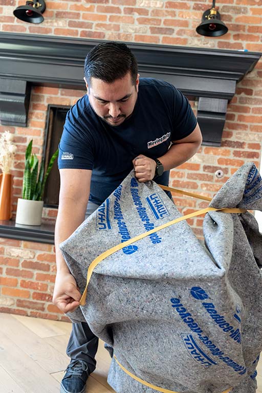 A Moving Helper carefully wraps a chair in furniture blankets and bands in a customer’s living room.