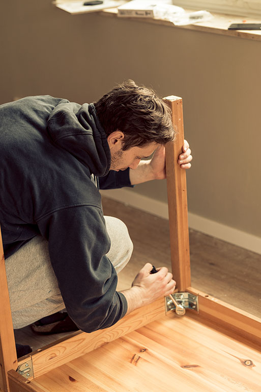 A man works to disassemble a wooden leg on his table. The answer to the question, “Do movers disassemble furniture,” is yes, most moving labor teams disassemble furniture.