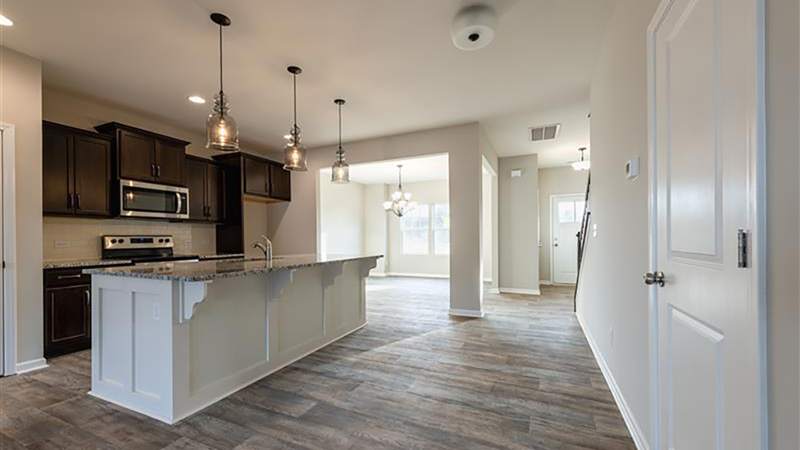 A modern kitchen and dining area convey elegance with hardwood floors, pendant lights over a granite island, dark cabinets, and a chandelier in the background.