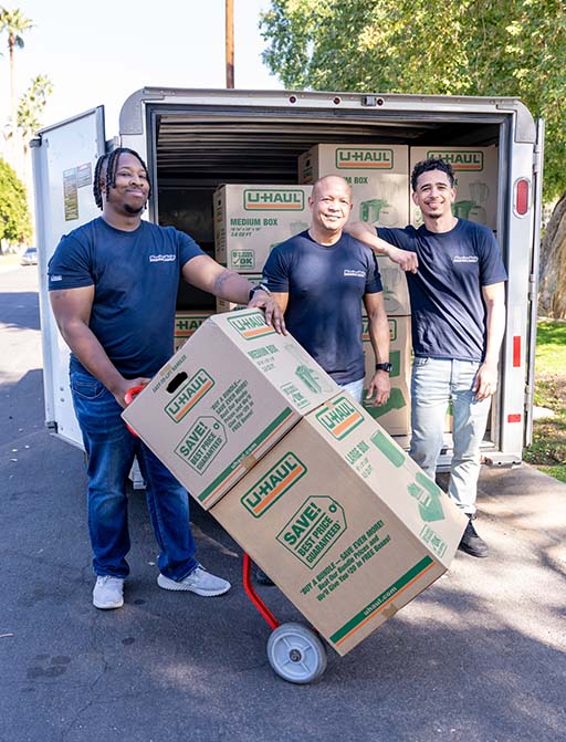 Three Moving Helpers smile together while one Moving Helper holds a utility dolly with moving boxes on it.