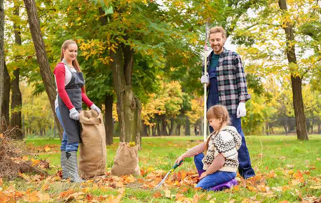 A father and his two daughters — one teenager and one child — work together to gather leaves in their backyard to clean their yard.