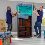 Two people wrapping a tall bookshelf in moving blankets and plastic stretch wrap. U-Haul plastic moving boxes are nearby in a bright, tidy room with large windows.