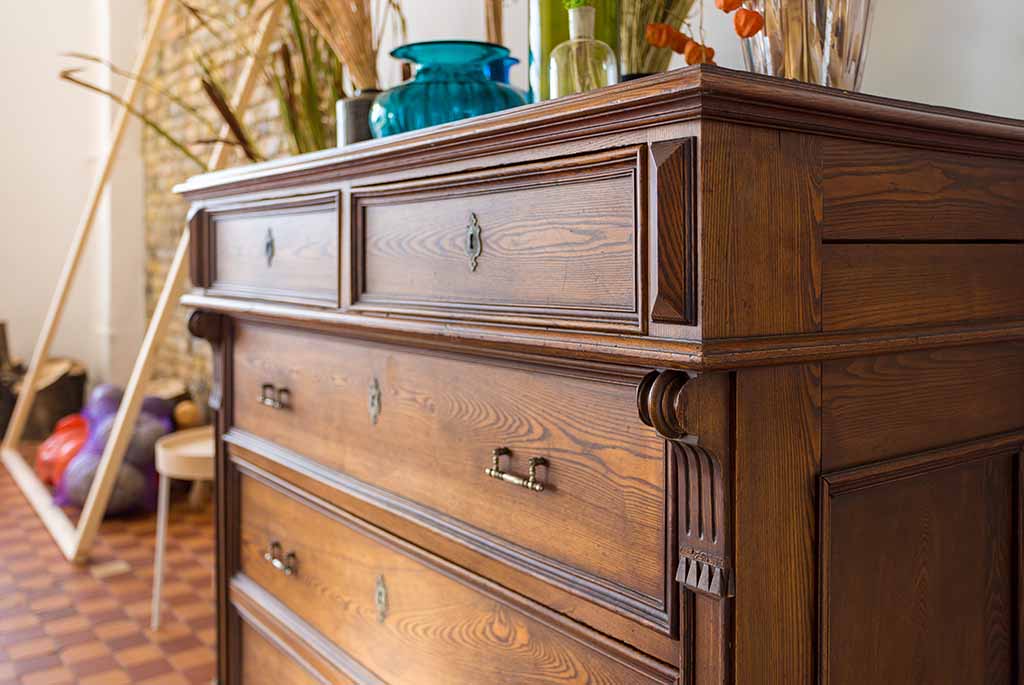 A close-up of an elegant wooden dresser with detailed carvings is shown in a bedroom. It features three drawers with metal handles, topped with decorative vases and plants.