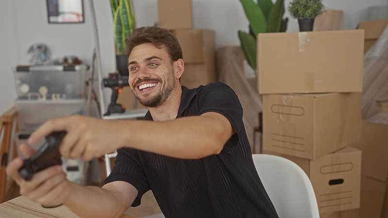 A man plays a game with his controller while moving boxes are stack around him in his new home.