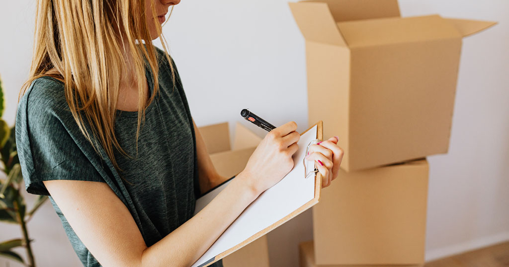 A woman writes something on her clipboard as she stands next to some opened moving boxes.