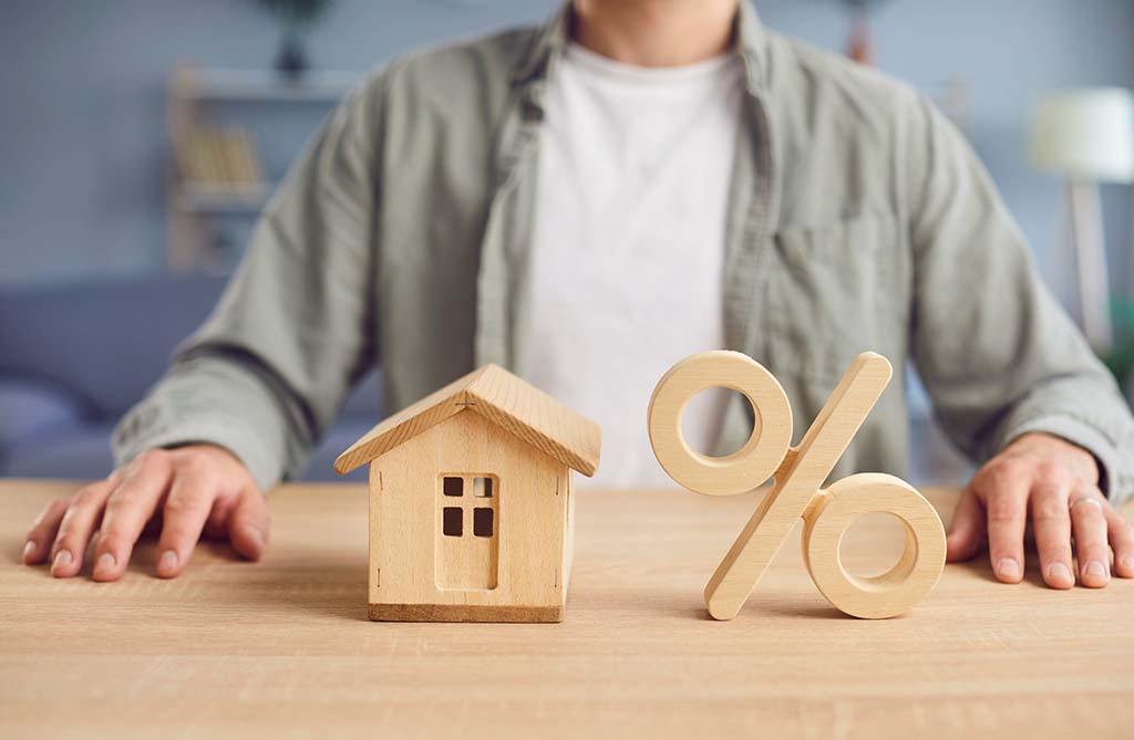 A man sits at a desk. A wooden house and a wooden percent sign rest on the table between the man’s hands.