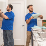 One Moving Helper carefully puts a glass cup into a cushion pouch before placing it into a moving box. Another Moving Helper puts a plate into a cushion pouch to put it into a different moving box.