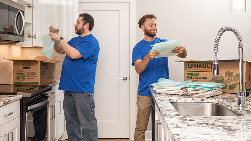 One Moving Helper carefully puts a glass cup into a cushion pouch before placing it into a moving box. Another Moving Helper puts a plate into a cushion pouch to put it into a different moving box.