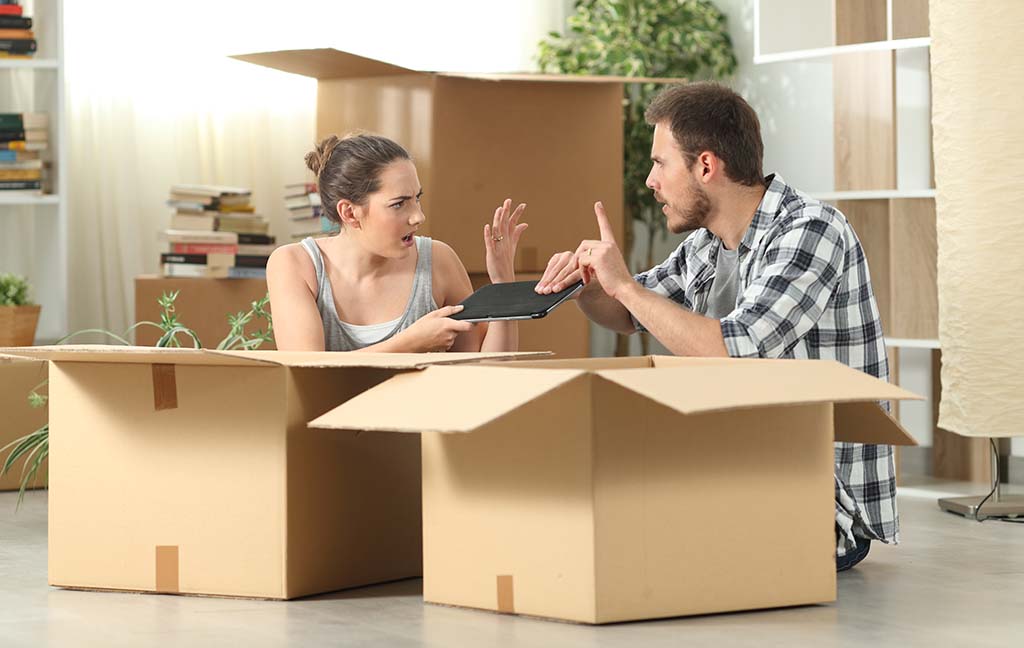 A couple sitting on the floor in front of two moving boxes argue over who gets to keep the tablet with each person grabbing it.