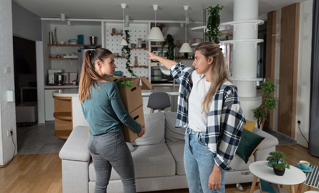 A woman points with her finger showing her ex-partner the door. The other woman carries two moving boxes while moving out after a breakup.