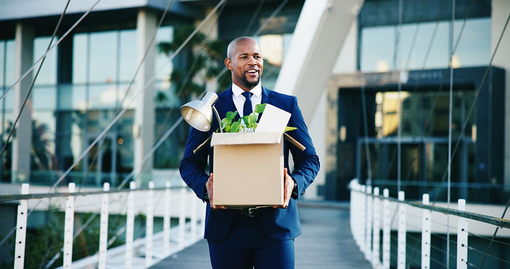 A man carries a box filled with work items from his desk as he leaves his old job’s building. Job relocation assistance may be available for your move.