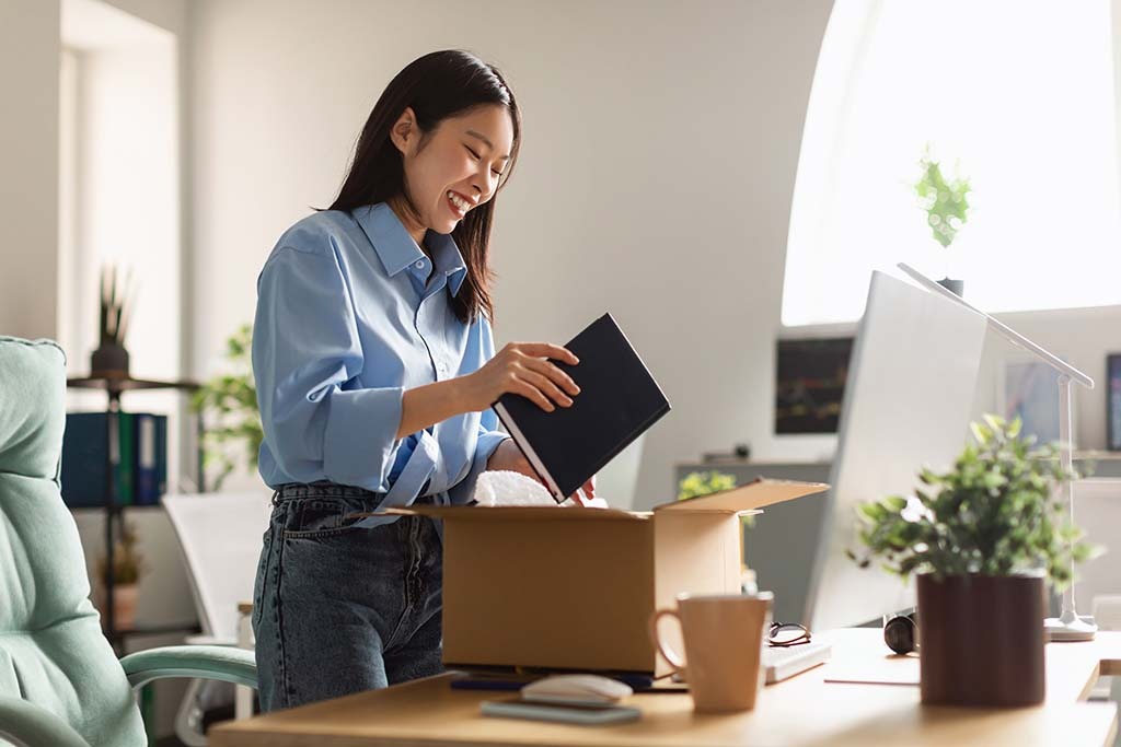 A woman places a book into a box as she packs her belongings from her desk on her last day at work. You can find affordable relocation services with the Moving Help Marketplace.