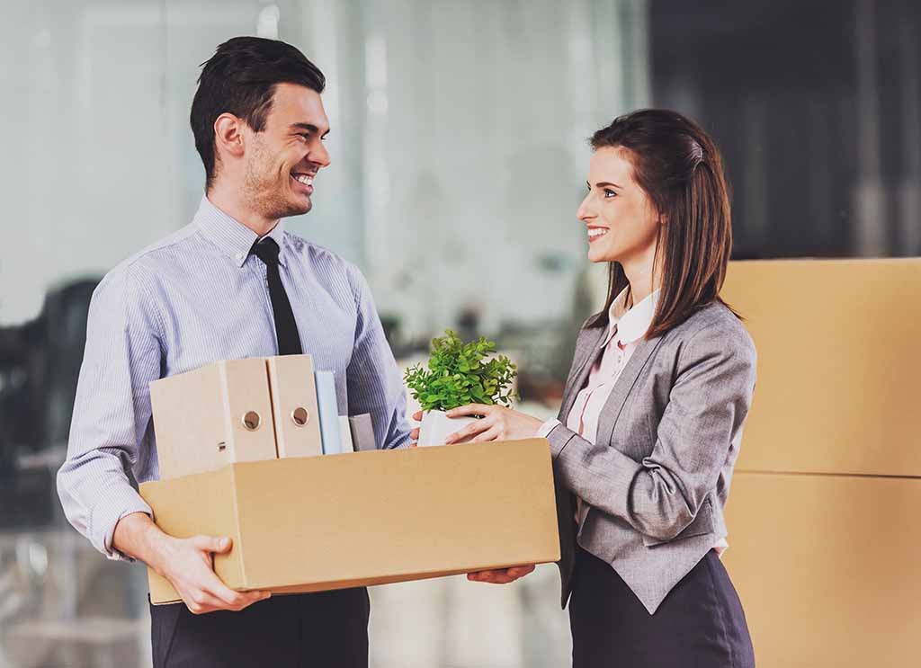 A man smiles back at his female colleague as she hands him his plant to place in the box he’s carrying. Some corporations offer a relocation package for new employees.