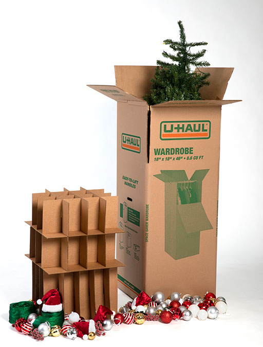 A Christmas tree is packed in a U-Haul Wardrobe Box. A U-Haul Cell Kit sits nearby with holiday ornaments placed around it on the ground.