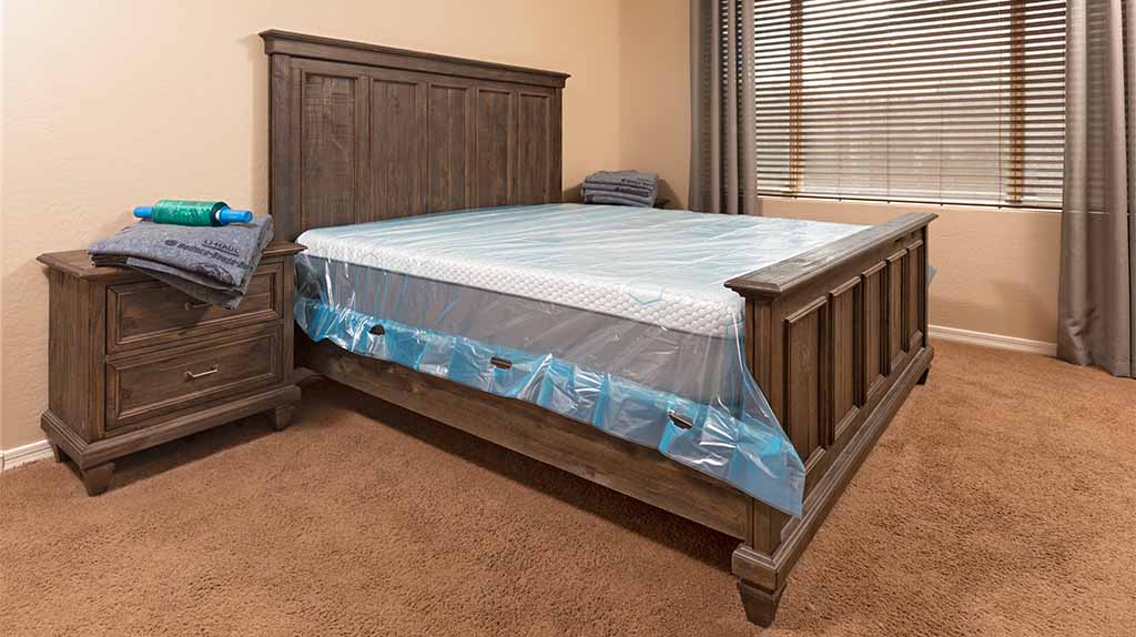 A U-Haul mattress pad covers a mattress in a bedroom. Next to the bed are two dressers with furniture blankets and mover’s plastic stretch wrap. It’s possible to figure out how to rent after an eviction.